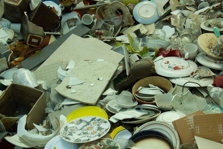 A large pile of assorted broken and discarded household waste and crockery, including shattered ceramic plates with floral patterns, smashed glassware, and various damaged kitchen utensils, all spread across a flat surface likely outdoors or on a paved area. Among the debris are broken bowls, cups, mugs, and fragments of plates in different sizes, shapes, and finishes, some with visible cracks or missing pieces. There are also pieces of damaged furniture or countertop materials, such as a large, dusty, white rectangular slab with chipped edges. The waste appears haphazardly piled, with some items leaning against each other, creating an environment that suggests an active rubbish clearance or an off-site collection of builders' waste. The scene has a neutral natural lighting with no visible background or environmental context, emphasizing the chaotic assortment of discarded domestic items. This type of clutter is typical of private waste handling or alternative disposal methods undertaken by services like Rubbish Clearance Mayfair for efficient rubbish removal or site clearance in areas with high foot traffic or renovation waste.