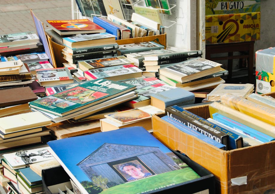 A large collection of books is spread across multiple wooden tables and in cardboard boxes, situated outdoors on a paved surface. The books vary in size, cover design, and thickness, with some stacked horizontally and others placed vertically, showcasing colorful and monochrome covers, including magazines, paperbacks, and hardcover editions. The textures range from glossy magazine pages to matte book covers, with some books leaning against each other or small stacks appearing slightly disorganized. In the background, a metal shelving unit with additional books and packaging is visible, along with a partially visible cardboard box containing more literature. The lighting appears natural, suggesting an outdoor or market environment, with no visible vehicle or machinery. This scene exemplifies a typical outdoor book sale or removal setup, where various printed materials are prepared for collection or disposal, aligning with private waste handling services like those provided by Rubbish Clearance Mayfair, specializing in efficient waste removal and clearance of non-standard items such as books or similar printed media.