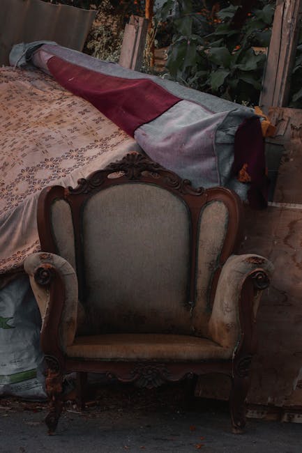 A vintage armchair with wooden carved details and beige fabric upholstery is positioned in a cluttered indoor or shed-like environment. The armchair has curved armrests and a high backrest, with a smooth, polished wooden frame. Behind the chair, there is a large rolled-up blanket or fabric covering, featuring a color-block design with sections of burgundy, gray, and light gray material, partially draped over a surface that appears to be a piece of furniture or a covered object. The surrounding area includes wooden panels or fencing, some lush green foliage visible to the right, and a textured floor surface. The scene is lit with natural, slightly subdued lighting, emphasizing the textures of the wooden frame and upholstery, and evoking an impression of temporary storage or a space used for miscellaneous household items that may be slated for disposal or removal as part of a rubbish clearance service by Rubbish Clearance Mayfair.