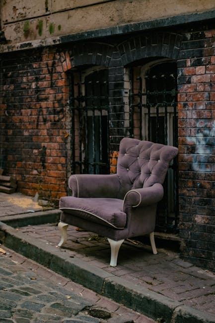 A plush, upholstered armchair with a tufted backrest and rolled armrests, covered in a muted purple-grey fabric, is positioned on a narrow sidewalk in front of a weathered brick building. The armchair features a textured, velvety surface and stands on four white-painted wooden legs with curved supports. Behind the chair, there are two black metal security bars covering tall, narrow windows set into the brick wall, which is aged with visible dark stains, soot marks, and patches of wear. The brickwork displays a mix of reddish and dark brown tones, with some areas of moss or dirt accumulation. The sidewalk consists of brick paving, slightly uneven, with a small curb separating it from the cobbled street. The environmental lighting suggests an overcast or shaded setting, typical of urban areas where private waste collection or alternative rubbish removal services, such as Rubbish Clearance Mayfair, might handle the removal of bulky furniture items like this armchair. Overall, the scene conveys an outdoor urban environment prepared for waste clearance on a street-side, aligning with services for private rubbish disposal outside conventional council collection.