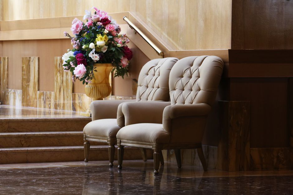 A vintage armchair with wooden carved details and beige fabric upholstery is positioned in a cluttered indoor or shed-like environment. The armchair has curved armrests and a high backrest, with a smooth, polished wooden frame. Behind the chair, there is a large rolled-up blanket or fabric covering, featuring a color-block design with sections of burgundy, gray, and light gray material, partially draped over a surface that appears to be a piece of furniture or a covered object. The surrounding area includes wooden panels or fencing, some lush green foliage visible to the right, and a textured floor surface. The scene is lit with natural, slightly subdued lighting, emphasizing the textures of the wooden frame and upholstery, and evoking an impression of temporary storage or a space used for miscellaneous household items that may be slated for disposal or removal as part of a rubbish clearance service by Rubbish Clearance Mayfair.