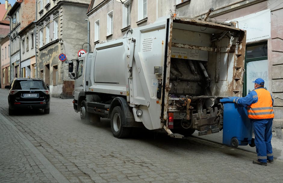 A street scene featuring a white waste collection vehicle parked beside a curb on a cobblestone pavement, with a worker dressed in blue overalls and an orange high-visibility vest loading a blue wheeled bin into the rear of the truck. The vehicle's rear compartment, designed for rubbish collection, has visible signs of rust and wear, indicating ongoing use for waste disposal. To the left of the waste truck, a black car is parked, and the surrounding area includes tall, multi-story buildings with a mixture of aged facades and modern elements, some featuring window shutters and exterior wall textures. The environment is illuminated by natural daylight, and the scene appears to be part of a private waste removal operation, possibly an independent collection service suitable for tailored rubbish removal that avoids relying solely on local authority collections. The overall setting emphasizes a typical urban waste collection process with a focus on efficient material handling and environmental hygiene in a city street context.