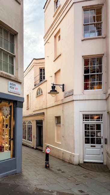 A wet, city street scene in Mayfair shows a corner with red-brick residential and commercial buildings featuring white window frames and decorative architectural elements. The pavement and road surfaces are reflective due to recent rain, with puddles visible on the sidewalk and the street. A pair of leafless trees with dark, slender branches stand in front of the buildings, contrasting against the overcast, grey sky. The street is empty of vehicles and pedestrians, with a slight incline leading around the corner to the right. The scene evokes an atmosphere of quiet urban life, where independent waste collection or private rubbish removal, such as those offered by Rubbish Clearance Mayfair, might be arranged as part of an on-site clearance or alternative waste handling service for clutter or antique clearing in the area.