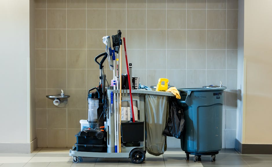 In a clean indoor setting, a mobile cleaning trolley loaded with various tools stands beside a large, gray waste bin on wheels. The trolley holds items including a black vacuum cleaner, a white bottle, a red mop, a blue mop, and other cleaning accessories, arranged in a compact manner. To the left of the trolley, a small wall-mounted stainless steel sink is visible, attached to a beige tiled wall. The environment appears orderly, with neutral-toned flooring and tiled walls, indicative of a communal or service area. The presence of the cleaning equipment and waste bin suggests activities related to cleaning and waste management, aligning with professional rubbish removal or on-site clearance services often used by businesses or facilities seeking private disposal solutions, rather than relying solely on municipal collection. Overall, the scene reflects an organized workspace with equipment essential for maintaining hygiene and managing waste efficiently, consistent with practices supported by companies like Rubbish Clearance Mayfair specializing in commercial waste duties.