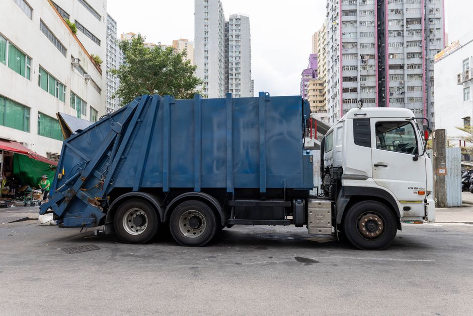 A large waste collection truck parked on an urban street, featuring a white cab and a blue, enclosed waste container with vertical reinforcement beams. The container appears to be made of metal with a smooth finish, and the rear section is angled downward, indicating its functionality for waste loading. The truck has six visible wheels, with the back dual wheels appearing slightly worn, and some rust visible on the underside and wheel rims. The vehicle is positioned on a paved road in front of multistory residential buildings with various coloured sections, including purple, pink, and beige facades, and some windows with green or brown frames. To the left, there are small street-side market stalls covered with green and red tarpaulins, and trees providing some greenery in the background. The overall scene suggests a typical city environment where commercial or residential waste is managed through independent collection services, reflecting the importance of private disposal options for urban waste management in Mayfair and similar areas.