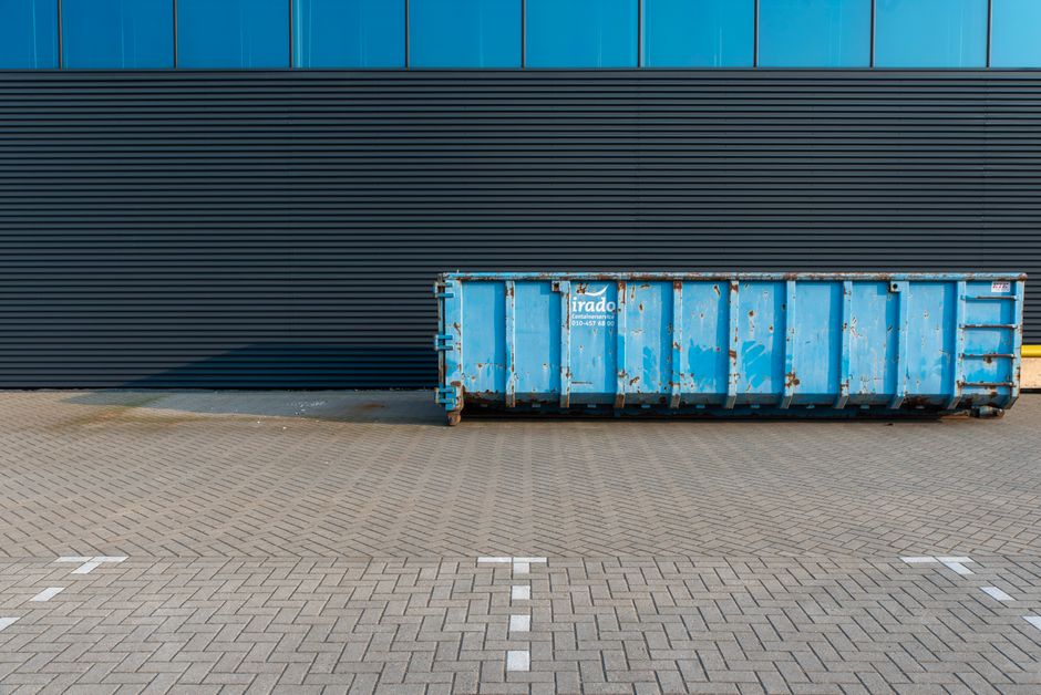 In a clean indoor setting, a mobile cleaning trolley loaded with various tools stands beside a large, gray waste bin on wheels. The trolley holds items including a black vacuum cleaner, a white bottle, a red mop, a blue mop, and other cleaning accessories, arranged in a compact manner. To the left of the trolley, a small wall-mounted stainless steel sink is visible, attached to a beige tiled wall. The environment appears orderly, with neutral-toned flooring and tiled walls, indicative of a communal or service area. The presence of the cleaning equipment and waste bin suggests activities related to cleaning and waste management, aligning with professional rubbish removal or on-site clearance services often used by businesses or facilities seeking private disposal solutions, rather than relying solely on municipal collection. Overall, the scene reflects an organized workspace with equipment essential for maintaining hygiene and managing waste efficiently, consistent with practices supported by companies like Rubbish Clearance Mayfair specializing in commercial waste duties.