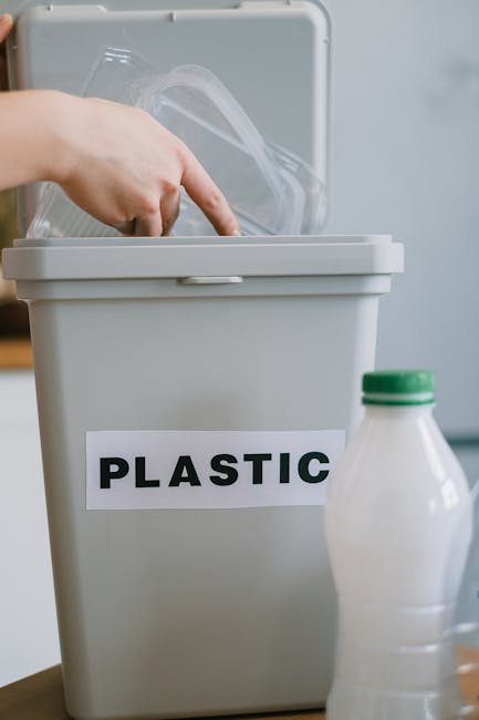 A close-up view of a grey plastic rubbish bin with a white label reading 'PLASTIC' in bold black letters, positioned on a wooden surface. Inside the open lid of the bin, a person's hand is seen placing a transparent plastic container with a clear lid. To the right of the bin, there is a partially visible white plastic bottle with a green cap. The background features a plain, neutral-colored wall, indicating an indoor environment. The image captures the act of separating plastic waste for disposal or recycling, highlighting the importance of organized rubbish sorting for effective waste management, with Rubbish Clearance Mayfair offering private waste collection services. The scene emphasizes environmentally conscious household waste handling and the practical aspect of on-site or independent collection solutions.