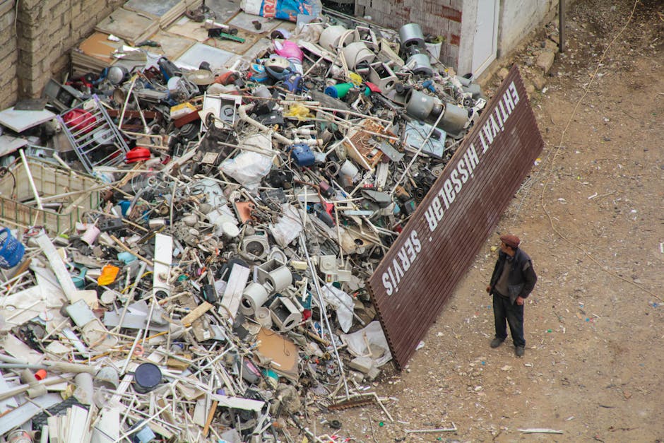 A large pile of mixed household waste and discarded items, including plastic, metal, and electronic debris, is situated on a construction site or outdoor area with a dirt ground. The trash consists of chairs, containers, broken appliances, and various miscellaneous household objects with differing materials and textures such as plastic, metal, and fabric. The pile is unorganized, overflowing from a nearby brick wall and partially obstructed by a brown metal sheet positioned at an angle, bearing the partially visible text 'THE WASHER SE...'. A man wearing a dark coat and brown hat stands to the right of the debris, observing the scene, in an environment lacking vegetation or paved surfaces, suggesting an open outdoor setting. The scene appears to relate to the context of private waste collection or on-site clearance, typical of rubbish removal services like those offered by Rubbish Clearance Mayfair, with the focus on collecting and managing household waste outside of municipal disposal systems.