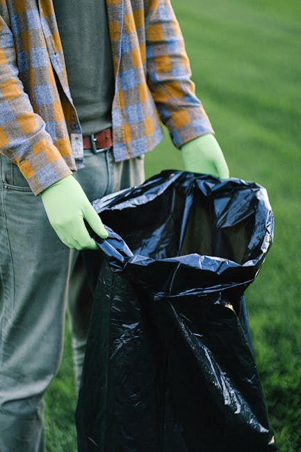 A close-up view of a grey plastic rubbish bin with a white label reading 'PLASTIC' in bold black letters, positioned on a wooden surface. Inside the open lid of the bin, a person's hand is seen placing a transparent plastic container with a clear lid. To the right of the bin, there is a partially visible white plastic bottle with a green cap. The background features a plain, neutral-colored wall, indicating an indoor environment. The image captures the act of separating plastic waste for disposal or recycling, highlighting the importance of organized rubbish sorting for effective waste management, with Rubbish Clearance Mayfair offering private waste collection services. The scene emphasizes environmentally conscious household waste handling and the practical aspect of on-site or independent collection solutions.