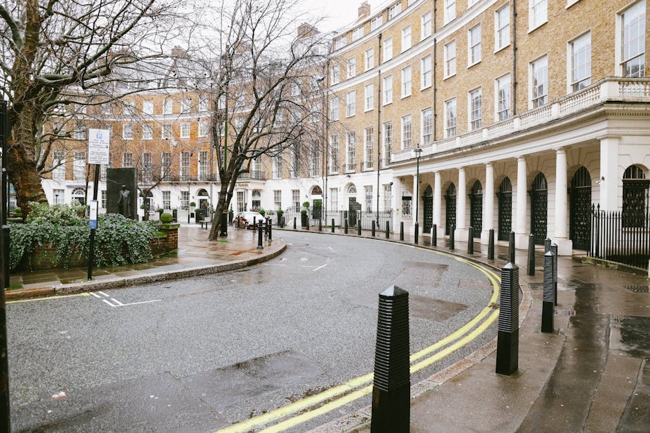 The image depicts a quiet street scene in Grosvenor Square with a curved asphalt road bordered by black plastic bollards and a double yellow line running along the edge. On the left side, a small paved area features a leafless tree with textured, grayish-brown bark and a bushy shrub with green foliage. There are several parked cars in the background, parked along the curb near elegant multi-storey Georgian-style buildings with light-colored brick facades, white window frames, and decorative stonework. The buildings have ground-floor entrances with black iron gates, and some windows are topped with small arched lintels. The pavement appears wet, reflecting a damp atmosphere, and soft, diffuse daylight filters through a cloudy sky. The overall scene is neat and orderly, characteristic of an affluent London neighbourhood, with subtle indications of private property and independent rubbish collection, as suggested by the absence of public waste bins or recycling facilities. Rubbish Clearance Mayfair's services are reflected in the context of discreet, on-site waste handling and clearance of residential or commercial refuse. The environment is calm, with no immediate activity, and the setting emphasizes the clean, well-maintained appearance of the street and surrounding buildings.