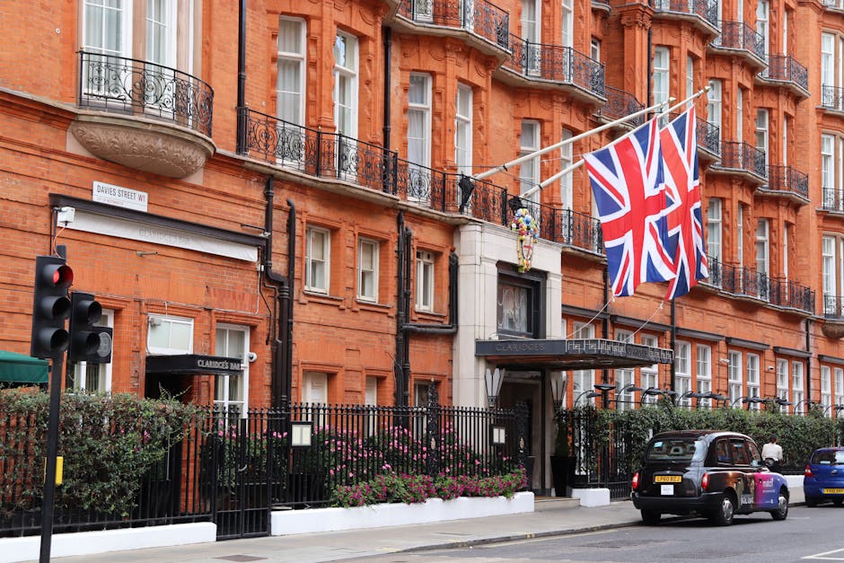The image shows a Victorian-style red brick building with ornate black wrought iron balconies and white-framed windows. A large Union Jack flag is prominently displayed, attached to a flagpole extending from the upper part of the building. At street level, there is a black metal fence surrounding a small garden area with neatly trimmed bushes and blooming pink flowers. A black entrance canopy with the words 'Claridges Hotel' is visible above the main door, which is situated within a cream-colored stone surround. To the left, a traffic light displaying a red signal is mounted on a pole beside the building. Several vehicles, including a black taxi and a small blue car, are parked along the street, which is paved with concrete. The environment appears bright with natural daylight, emphasizing the building's warm tones and the flags fluttering in a gentle breeze, suggesting a busy urban setting where private collection or on-site clearance services, such as those provided by Rubbish Clearance Mayfair, might be needed for waste and rubbish removal in this upscale area.