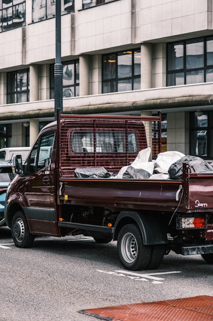 A small burgundy flatbed truck parked on an urban street in front of a modern, multi-story building with large glass windows and a concrete facade. The truck's bed is loaded with a collection of mixed waste, including black, white, and grey bags, some of which are partially open, revealing miscellaneous debris and materials inside. The truck features a metal safety grille behind the cab and silver hubcaps on its wheels, with a noticeable side mirror extending from the driver's side. The street surface is paved, with a white line and a red tactile paving strip visible at the bottom edge of the image. A tall, dark streetlight pole stands adjacent to the vehicle, and neighboring cars and buildings indicate a busy city environment. This scene illustrates a private waste collection service, relevant to companies like Rubbish Clearance Mayfair, providing an alternative to council rubbish disposal for commercial or residential rubbish removal in a metropolitan setting.