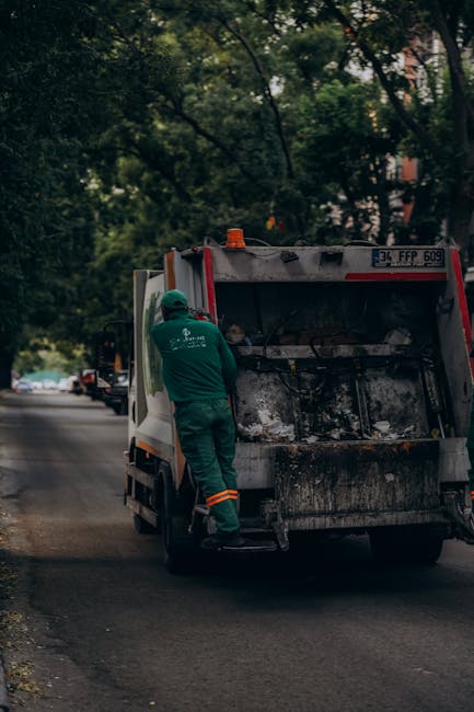 A worker dressed in green uniform and safety helmet is standing beside the open rear of a black garbage truck, which is parked on the side of a residential street with trees and parked cars visible in the background. The truck's loading area contains black waste material, likely rubble or general rubbish, with signs of dirt and soot. The worker appears to be engaging in waste collection or maintenance activity, consistent with private rubbish clearance services provided by Rubbish Clearance Mayfair. The scene is set during daylight hours, with natural ambient light highlighting the street environment and the utilitarian design of the vehicle, emphasizing the process of on-site waste collection in an urban setting, aligned with alternative waste handling practices outside of local authority disposal channels.