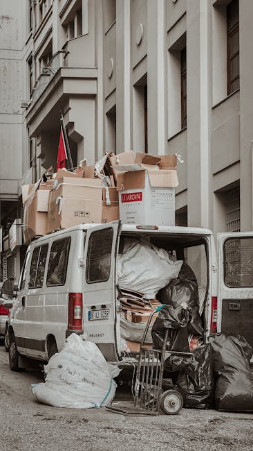 A small burgundy flatbed truck parked on an urban street in front of a modern, multi-story building with large glass windows and a concrete facade. The truck's bed is loaded with a collection of mixed waste, including black, white, and grey bags, some of which are partially open, revealing miscellaneous debris and materials inside. The truck features a metal safety grille behind the cab and silver hubcaps on its wheels, with a noticeable side mirror extending from the driver's side. The street surface is paved, with a white line and a red tactile paving strip visible at the bottom edge of the image. A tall, dark streetlight pole stands adjacent to the vehicle, and neighboring cars and buildings indicate a busy city environment. This scene illustrates a private waste collection service, relevant to companies like Rubbish Clearance Mayfair, providing an alternative to council rubbish disposal for commercial or residential rubbish removal in a metropolitan setting.