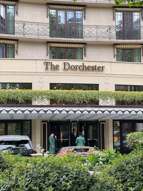 The entrance of The Dorchester hotel in Mayfair, featuring a beige stone facade with large glass doors and gold trim accents. The hotel name is displayed prominently in dark lettering above the entrance. In front of the building, there is a small landscaped area with neatly trimmed green bushes and flowering plants. To the left, a black luxury vehicle is parked close to the entrance, and three individuals dressed in formal attire are engaged in conversation near the doorway, suggesting a private or professional meeting. The upper floors of the building have large windows with black wrought-iron balcony railings on each level, and the overall scene is set during daylight with natural light illuminating the structure and surrounding greenery. This setting reflects a high-end London location, where waste removal services may include private collection options for hotel or commercial premises, as exemplified by Rubbish Clearance Mayfair's work handling waste in such environments.