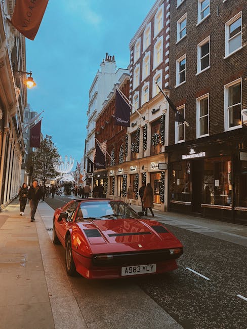 A red vintage sports car with a sleek, low-profile design and glossy finish is parked on the street, facing the camera. The vehicle features a distinctive angular shape, pop-up headlights, and a narrow front grille, situated close to the curb on the left side of the image. Behind the car, there are several pedestrians walking along the sidewalk, some entering or exiting shops and cafes housed in multi-storey buildings with a mix of modern and classic facades. The buildings on the right are constructed of brick and concrete, with large windows and dark-colored signage, and are lined with black flags displaying the brand name or logo. The street is illuminated by warm exterior lighting from the shopfronts and street lamps, contrasting with the bluish evening sky. In the background, a decorated city street is visible, with string lights hanging across the road, contributing to a lively yet orderly urban atmosphere. The scene appears to depict a vibrant city area potentially involved with private street parking or independent vehicle storage, consistent with the context of alternative waste handling and rubbish removal services provided by Rubbish Clearance Mayfair.