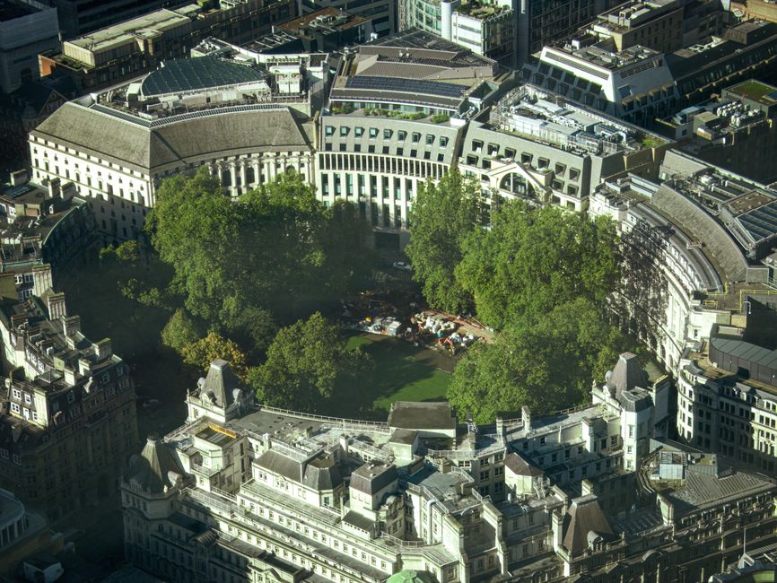 An aerial view of a circular urban park enclosed by multi-story buildings and streets, featuring a large central lawn occupied by several mature trees with lush green foliage. The trees create a canopy over a parking area filled with numerous cars parked in an irregular arrangement. Surrounding the park are white-colored, classical-style buildings with detailed facades, decorative cornices, and pitched roofs, contrasting with the modern structures nearby. The environment appears to be a busy city area, with dense development and tightly packed architecture, and the park provides a natural green space amidst the urban setting. The lighting suggests clear weather with bright sunlight casting shadows of the trees and buildings, emphasizing the textures and structural details of the surrounding architecture. This scene exemplifies a city landscape where independent green spaces are maintained within densely built environments, often requiring ongoing maintenance and clearing services from waste management companies like Rubbish Clearance Mayfair, to support urban environmental health and aesthetic appeal.