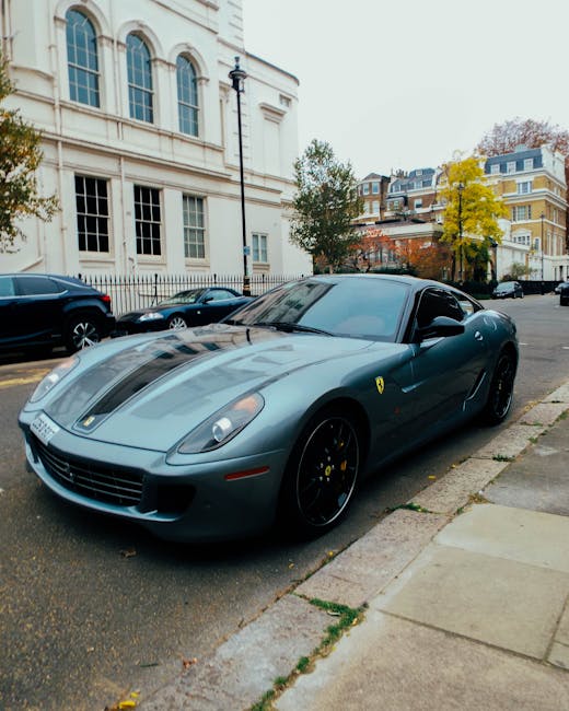 A red vintage sports car with a sleek, low-profile design and glossy finish is parked on the street, facing the camera. The vehicle features a distinctive angular shape, pop-up headlights, and a narrow front grille, situated close to the curb on the left side of the image. Behind the car, there are several pedestrians walking along the sidewalk, some entering or exiting shops and cafes housed in multi-storey buildings with a mix of modern and classic facades. The buildings on the right are constructed of brick and concrete, with large windows and dark-colored signage, and are lined with black flags displaying the brand name or logo. The street is illuminated by warm exterior lighting from the shopfronts and street lamps, contrasting with the bluish evening sky. In the background, a decorated city street is visible, with string lights hanging across the road, contributing to a lively yet orderly urban atmosphere. The scene appears to depict a vibrant city area potentially involved with private street parking or independent vehicle storage, consistent with the context of alternative waste handling and rubbish removal services provided by Rubbish Clearance Mayfair.