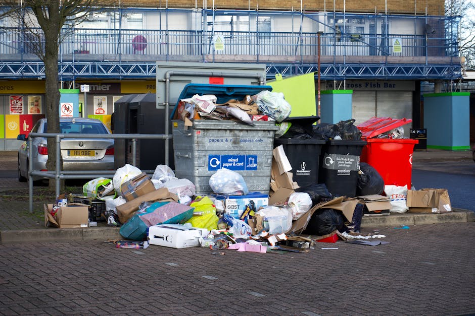 A collection of overflowing rubbish bins and scattered waste including cardboard boxes, plastic bags, paper, and packaging materials positioned on a paved area beside a metal railing. The waste appears haphazardly piled, with some bags black and others transparent, revealing mixed refuse inside. The bins include a large grey recycling bin labeled for mixed paper and cardboard, a black bin for general waste, and a prominent red bin, all situated near a curb in an urban setting. In the background, there is a multi-storey building under construction or renovation, covered with blue scaffolding and mesh netting, with a row of closed shopfronts on the ground level, including signage for a fish bar and other commercial units. A parked car is visible on the left. The scene is lit with natural daylight, casting soft shadows, illustrating a typical example of private waste accumulation awaiting removal by a rubbish clearance service such as Rubbish Clearance Mayfair, operating within the context of professional waste management and alternative disposal methods.
