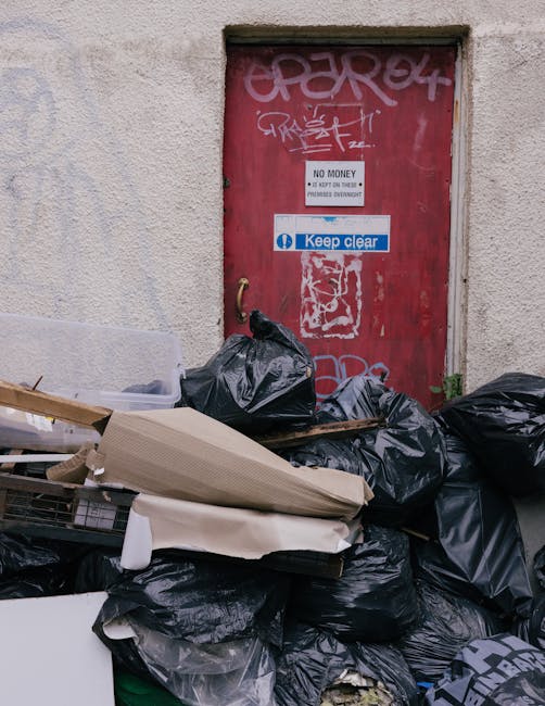 A collection of black and grey rubbish bags piled up against a rough, beige exterior wall below a red metal door covered in graffiti and stickers. The bags appear filled with various waste materials, some bulging and others tightly sealed, with a few loose debris and flattened cardboard boxes resting on top. The door is framed by a concrete surround and has a small vent or opening above it. Signs on the door include a white notice indicating 'No Money' and a blue 'Keep Clear' sticker, suggesting restricted access or private property. The scene reflects an outdoor rubbish accumulation, typical of an area awaiting collection or removal, with visible environmental details consistent with urban waste handling that private waste removal companies like Rubbish Clearance Mayfair could address through on-site clearance services.