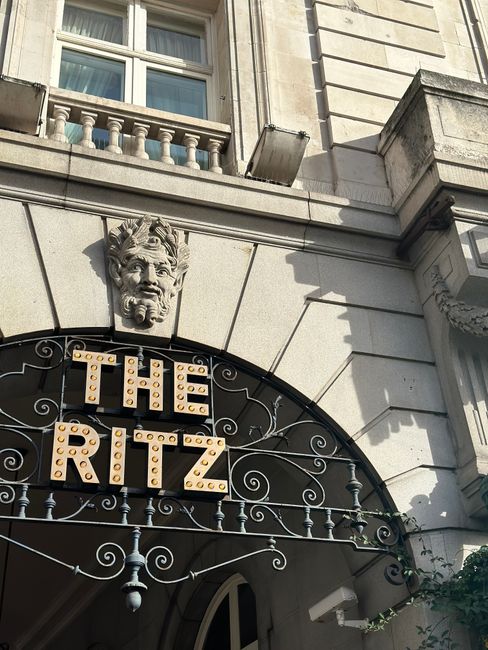 Close-up view of the exterior of a historic building showing ornate architectural details, including a decorative stone lion's head sculpture positioned above a rounded archway. Beneath the sculpture, there is a sign spelling 'THE RITZ' in bold, three-dimensional letters with rounded edges, illuminated by small light bulbs. The building's facade is constructed from light grey stone blocks, featuring finely carved horizontal lines and a textured surface. To the right of the sign, part of a window with a white frame and clear glass is visible, set into the room's wall and divided by horizontal and vertical muntins. An external metal canopy with a decorative wrought iron framework, painted black with subtle scrollwork, extends over the entrance, supported by central vertical rods and horizontal bars. The surrounding environment hints at a city street with natural daylight illuminating the scene, casting shadows on the building's facade. This detailed architectural style and signage are characteristic of a high-end or historic hotel, fitting within the context of independent waste collection and private disposal services for such establishments, as provided by Rubbish Clearance Mayfair.
